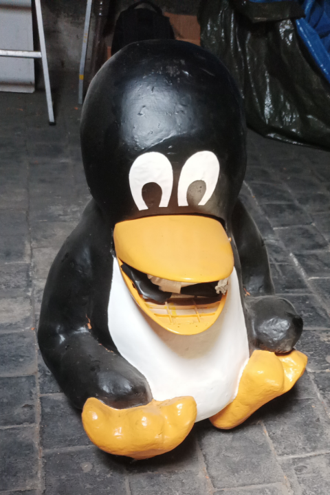 Un manchot en plâtre assis dans la pause iconique de Tux, se tenant la patte gauche. Son bec est un rack pour lecteur CD/DVD.